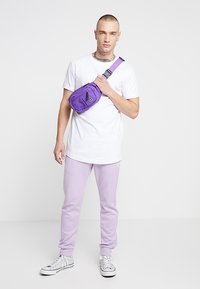 Lila Stoff-Crossbody-Tasche mit Reißverschluss-Tasche, getragen über einem weißen T-Shirt und lavendelfarbenen Jogginghosen. Die Sneaker sind weiß mit schwarzen Akzenten.