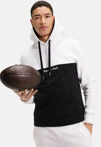 Sweatshirt noir et blanc à capuche, avec une poche kangourou, des cordons de serrage, et un logo sur la poitrine, tenu avec un ballon de rugby oval brun.