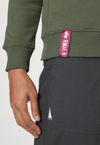 Sweatshirt verde oliva con polsini e orlo a coste. Presenta un'etichetta rosa con testo bianco. Abbinato a pantaloni tecnici leggeri di colore grigio scuro.