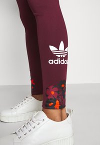 Maroon leggings with floral accents in red and black at the hem, featuring a large white Adidas logo on the side. Worn with white sneakers.