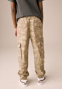 Pantaloni cargo mimetici beige con polsini elastici, tasche laterali con zip e tasche posteriori, abbinati a scarpe da ginnastica Nike bianche e marroni.