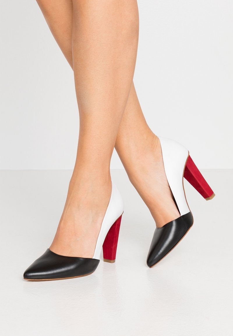 Jambes portant des chaussures à talons hauts avec des bouts pointus noirs, des côtés blancs et des talons rouges épais, debout sur une surface claire.