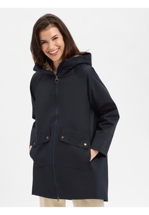 Vrouw glimlachen draagt een marineblauwe regenjas met capuchon, ritssluiting aan de voorkant en klepzakken, staand met handen in de zakken.