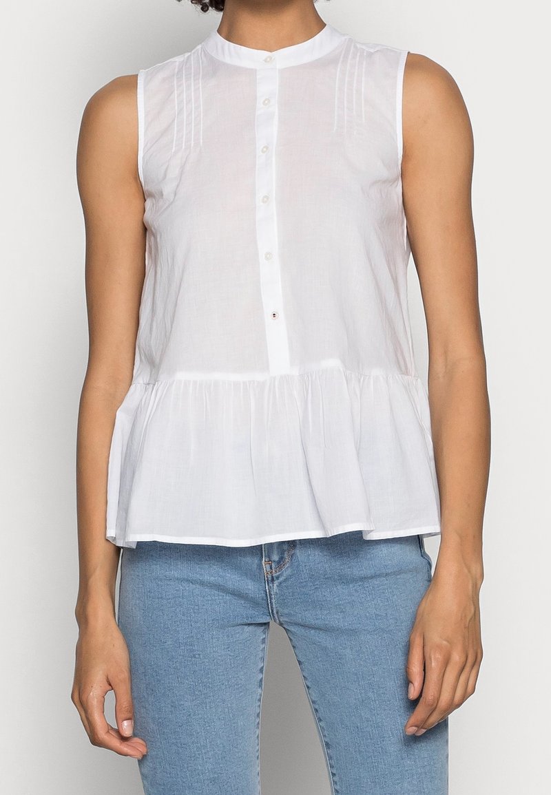 Blouse blanche sans manches avec un col rond, un devant à boutons et un détail de basque froncée à la taille. Tissu léger avec une texture subtile.