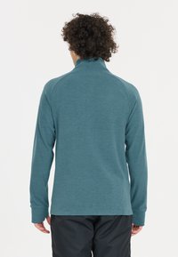 Teal pitkähihaisen poolopuseron, jossa on korkea kaulus, raglan-hihat ja teksturoitu kangas. Ei näkyviä metalliosia. Sileä pinta.