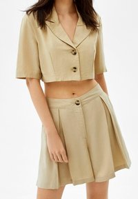 Beige kort blazer med korta ärmar och två knappar, matchat med högmidjade kavajshorts med en matchande knapp. Slät tygtextur.