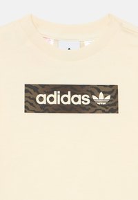 Camiseta de color crema con un logotipo rectangular de camuflaje oscuro de Adidas y el símbolo trefoil centrados debajo del cuello redondo acanalado.