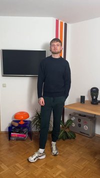 Pull bleu marine foncé, pantalon vert ajusté et baskets argentées. Cheveux courts et tatouages visibles. En arrière-plan, une télévision, une lampe et des plantes.