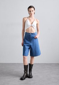 Weißes Halter-Crop-Top mit verdrehtem Detail, kombiniert mit blauen Denim-Bermuda-Shorts, ergänzt durch schwarze kniehohe Chunky-Stiefel.