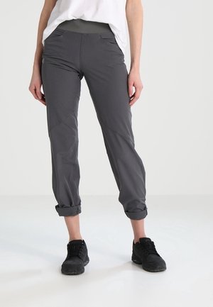 Person iført mørkegrå opsmøgede bukser, sorte sneakers og en hvid skjorte, der står på en lys gulv mod en ensfarvet baggrund.
