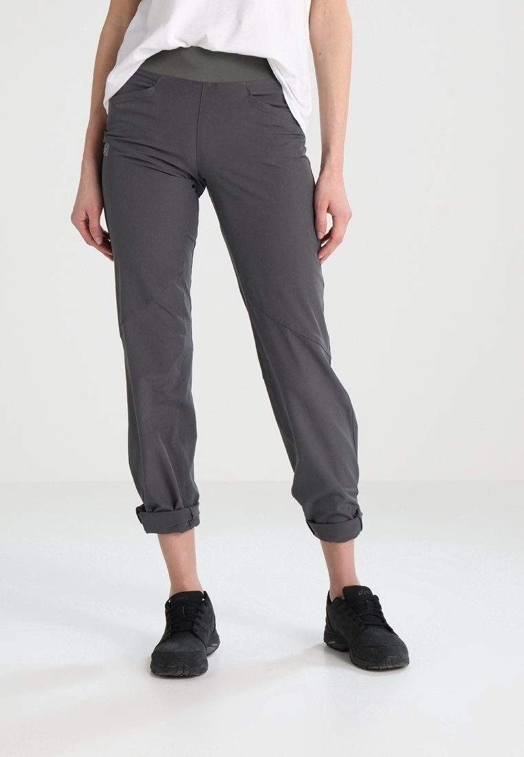 Personne portant un pantalon gris foncé retroussé, des baskets noires et une chemise blanche debout sur un sol clair devant un fond uni.