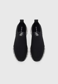 Sapatos pretos slip-on feitos de malha respirável com superfície texturizada, apresentando sola branca e detalhe de logo prateado na parte superior.