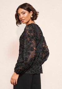 Črna čipkasta bluza z dvignjenim cvetličnim vzorcem, dolgimi napihnjenimi rokavi in okroglo izrezom. Tkanina je polprosojna z zapletenimi detajli.