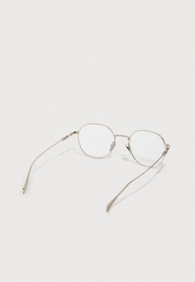 Armação de óculos de metal prateado com estrutura fina e delicada, lentes redondas e detalhes gravados nas hastes. Lentes transparentes.