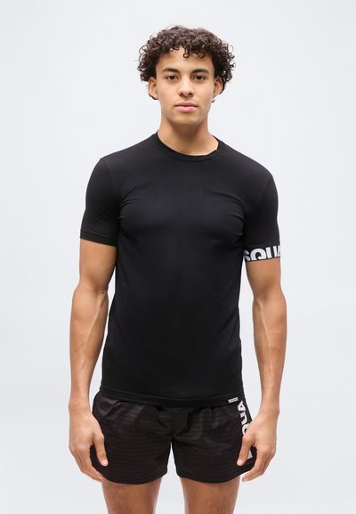 Jeune homme aux cheveux bouclés portant un T-shirt noir ajusté avec un texte blanc sur la manche et un short noir à motif subtil.