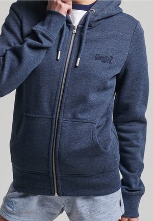 Osoba nosząca granatową bluzę z kapturem zapinaną na zamek z troczkami i jasnoszare spodenki, ręka blisko kołnierza, na jednolitym tle.