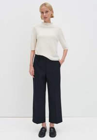 Donna in piedi con le mani nelle tasche di pantaloni larghi neri, indossa una maglia bianca a maniche corte con collo alto corto e mocassini neri, su sfondo semplice.