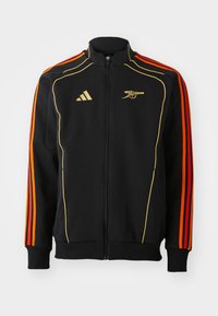Veste zippée noire avec des accents dorés et oranges. Présente trois rayures oranges sur les manches et un logo sur la poitrine. Fabriquée en tissu doux.