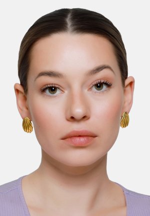 Giovane donna con capelli scuri tirati indietro, che indossa orecchini dorati a forma di conchiglia e una maglietta lavanda, espressione facciale neutra, sfondo uniforme.