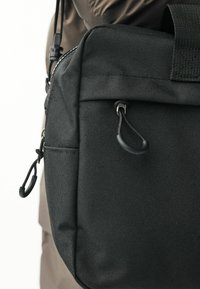 Zwarte stoffen tas met een gestructureerd oppervlak, voorzien van een rits, een klein vak en een trekkoordlus voor een veilige sluiting.