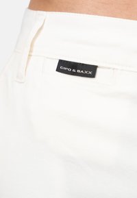 Tissu en denim blanc en coton avec une poche arrière visible. Présente une étiquette noire avec le texte "CIPO & BAXX" et une bordure texturée et cousue.