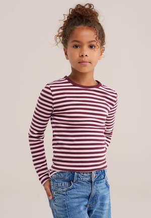 Une jeune fille aux cheveux bouclés pose devant un fond uni. Elle porte un haut à manches longues rayé bordeaux et blanc ainsi qu’un jean bleu.