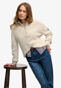 Femme s'appuyant sur un tabouret en bois, vêtue d'un pull en maille crème sur une chemise rayée et un jean bleu, regardant sur le côté.