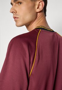 Bordeaux sportshirt van een gestructureerde stof, met een zwarte en gele kraag en zijaccenten, en een ronde halslijn.