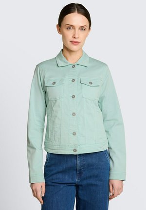 Kobieta ubrana w jasną, miętowozieloną kurtkę dżinsową zapinaną na guziki oraz niebieskie jeansy, stojąca na tle jednolitego jasnego tła.