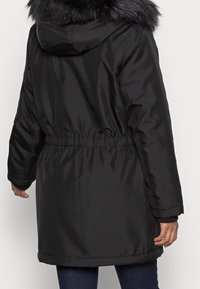 Musta toppatakki, jossa on karvavuorattu huppu, joustava vyötärö, pitkät hihat ja hieman kaareva helma. Sileä pinta, käytännöllinen ja lämmin malli.