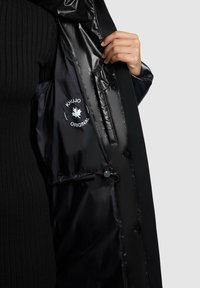 Chaqueta acolchada negra con un forro interior brillante, que presenta un parche con el logo, botones a presión y un detalle de cordón para un ajuste ajustable.