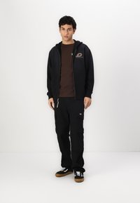 Črna zip-up kapuca z logotipom, rjav t-shirt in črne cargo hlače; črne čevlje z belimi detajli in rjavimi podplati. Preprost, funkcionalen dizajn.