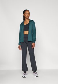 Giacca zip-up verde scuro sopra un crop top nero, abbinata a pantaloni neri a vita alta e sneakers bianche con dettagli rosa.