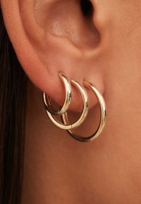 Boucles d'oreilles en or à trois niveaux mettant en valeur une finition polie. Chaque anneau a une forme lisse et arrondie, posée sur une oreille de teinte peau.