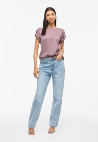 Light mauve short-sleeve top with rolled cuffs paired with high-waisted, loose-fitting light blue jeans; model stands with hands on hips.