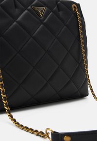 Czarna pikowana torebka o błyszczącym wykończeniu, z złotym emblematem i łańcuszkowym paskiem. Charakterystyczny diamentowy wzór i gładka tekstura.