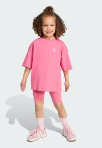 Lyserød t-shirt med korte ærmer, parret med lyserøde cykelshorts. Hvid Adidas-logo på t-shirten. Fodtøjet inkluderer lyserøde sneakers med hvide detaljer.