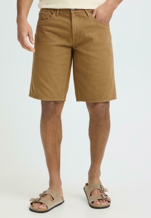 Mężczyzna w brązowych krótkich spodenkach do kolan i beżowych sandałach z odkrytymi palcami, stojący z jedną ręką w kieszeni na jasnym tle.