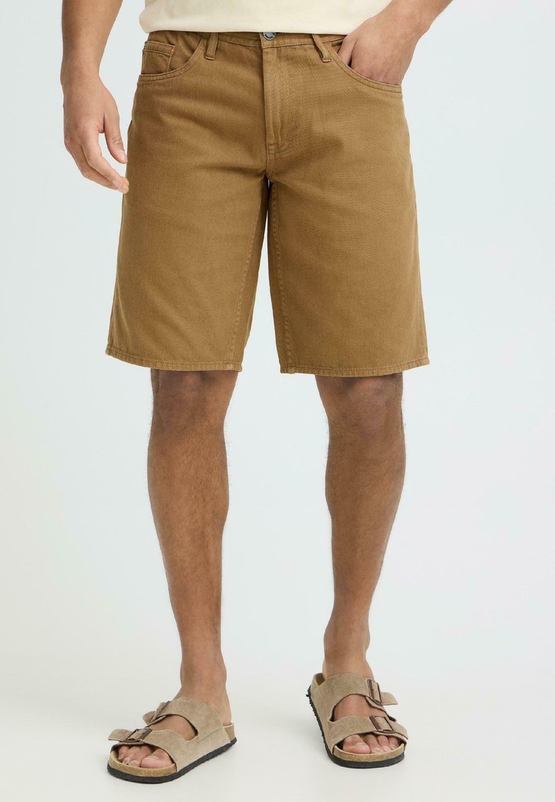 Άνδρας με καφέ σορτς μέχρι το γόνατο και μπεζ σανδάλια ανοιχτού τύπου, στέκεται με το ένα χέρι στην τσέπη σε ανοιχτό φωτεινό φόντο.