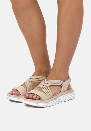 Füße in beigen sportlichen Sandalen mit überkreuzten Riemen und dicken weißen Sohlen vor weißem Hintergrund.