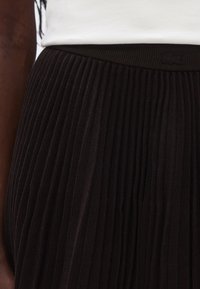 Jupe plissée noire avec une ceinture côtelée. Le tissu a une texture lisse, présentant de fines plis verticaux et un accent logo.