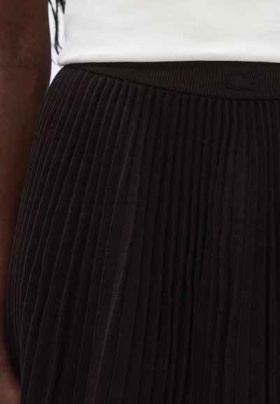 Jupe plissée noire avec une ceinture côtelée. Le tissu a une texture lisse, présentant de fines plis verticaux et un accent logo.