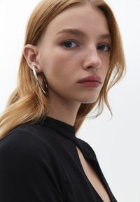 Stříbrné kruhové náušnice, piercing v septu, hladká pleť, dlouhé vlnité blond vlasy, černé tričko s vysokým výstřihem, neutrální pozadí.