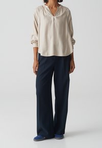 Blusa leggera beige con scollatura a V e maniche lunghe, abbinata a pantaloni larghi blu navy. Scarpe blu completano l'outfit. Texture morbida, vestibilità comoda.