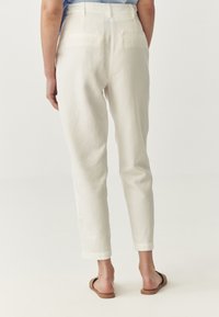 Pantaloni bianchi di lino con gamba affusolata, due tasche posteriori e orli risvoltati, abbinati a sandali scivolati marroni. Texture morbida e design leggero.