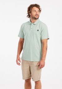 Lichtgroene polo met een kraag, drie knopen en een borstzak. Draag het met beige shorts. Gladde stoftextuur.