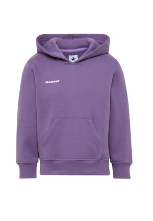 Fille aux cheveux bouclés portant un sweat à capuche violet, un pantalon vert avec des genoux renforcés, des baskets blanches et un sac à dos vert avec une petite peluche noire.