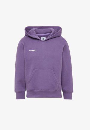 Fille aux cheveux bouclés portant un sweat à capuche violet, un pantalon vert avec des genoux renforcés, des baskets blanches et un sac à dos vert avec une petite peluche noire.