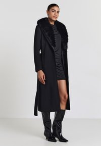 Cappotto lungo nero con colletto in pelliccia sintetica, caratterizzato da un taglio dritto, tasche laterali e una texture lucida. Indossato sopra un vestito scuro arricciato.