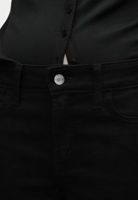 Κοντινή λήψη μαύρου τζιν ψηλόμεσου με ασημένιο κουμπί και θηλιές για ζώνη, συνδυασμένο με μαύρο ριπ τοπ με κουμπιά, που είναι μπουκλωμένο μέσα.